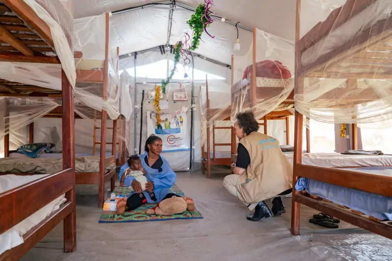 A caretaker at a nursery sits with a child in her lap speaking to a woman squating next to her.