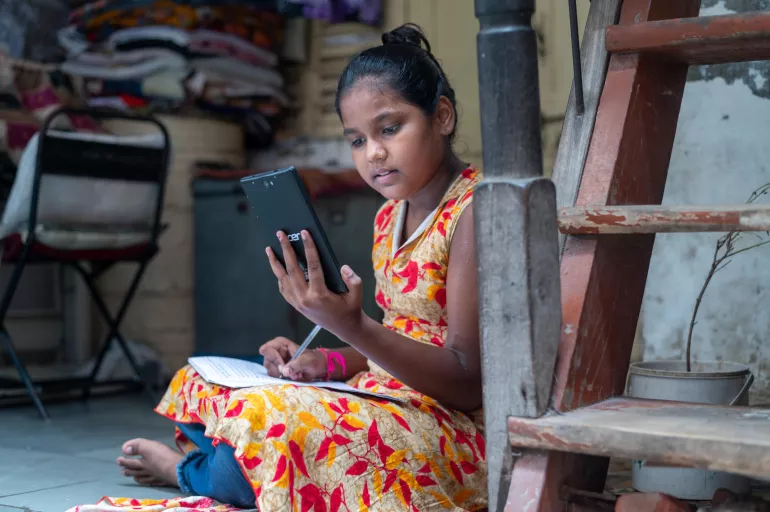 A girl child sits cross-legged on the floor holding a tablet and notebook.