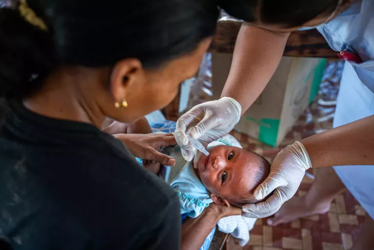 An infant is held by their mother while a healthcare provider vaccinates them with a mouth dropper.