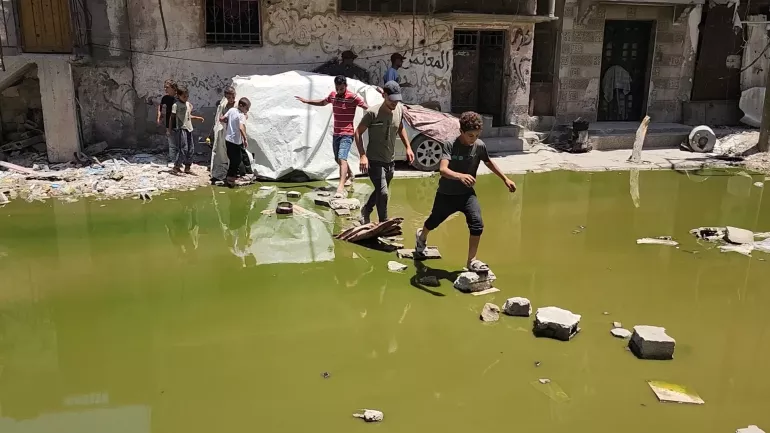 Children step across a series of stones to cross a pool of contaminated water. 