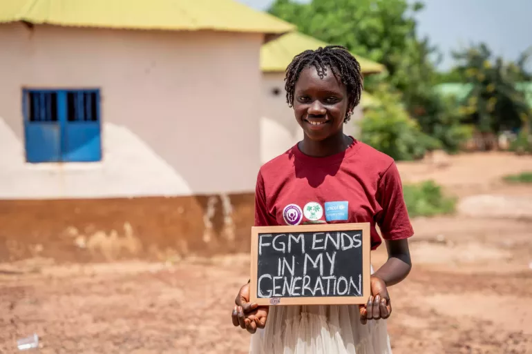 An adolescent girl holds a sign outside. The sign says "FGM ends in my generation."