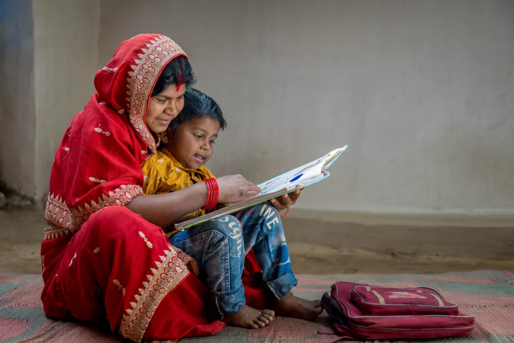 India: A woman wearing a red sari sits on the floor with her son in her lap reading a book together.