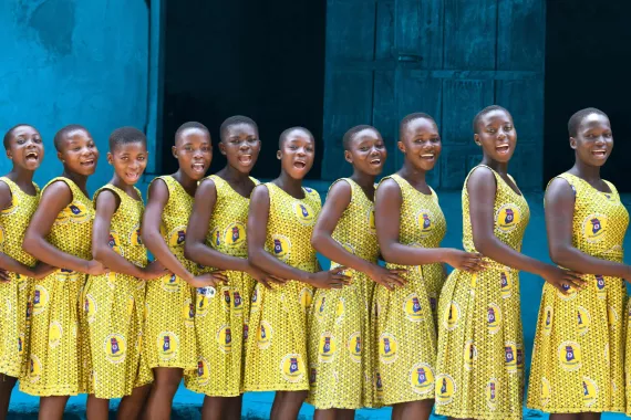 A group of schoolgirls in Ghana who are all wearing matching dresses stand in a line behind each other