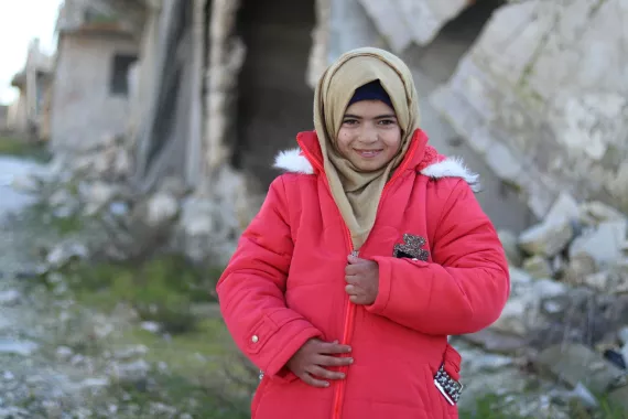 A young girl standing in a refugee camp wearing a bright pink winter coat
