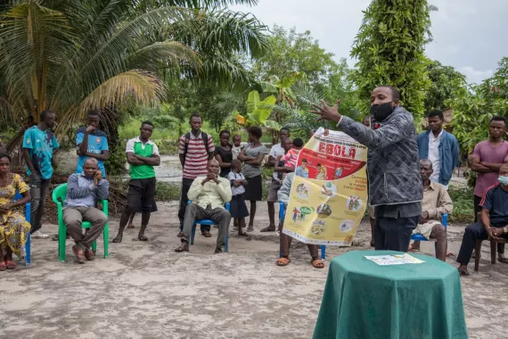 A man wearing a mask and holding a poster about Ebola talks to a group of community members who are sitting and standing in a circle around him