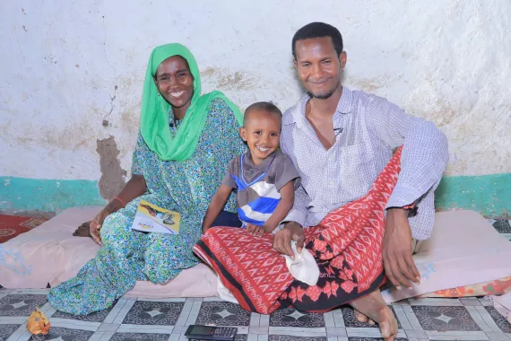 A mother, faher and their young child who are all similing, sitting on cushions on the ground.