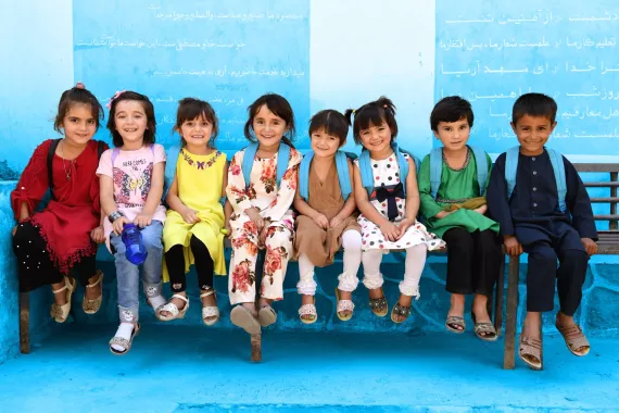A row of young smiling school students sitting on a long bench together with Arabic script on the wall behind them