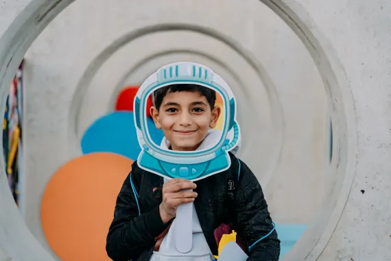 A boy holds an paper astronaut helmet in front of his face while smiling.