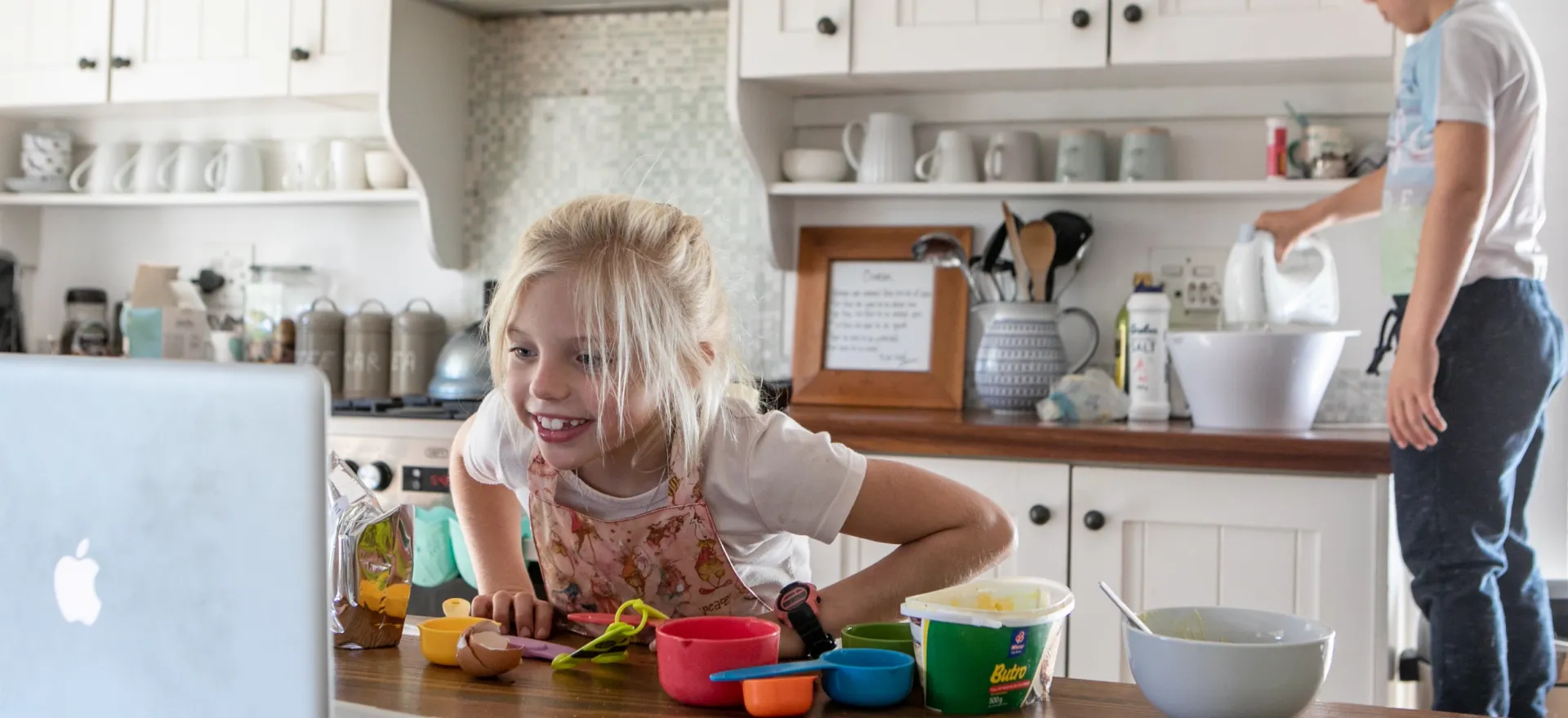 A girl leans on a kitchen counter to watch a video on a lap on baking, while her brother is standing on a chair while mixing ingredients.