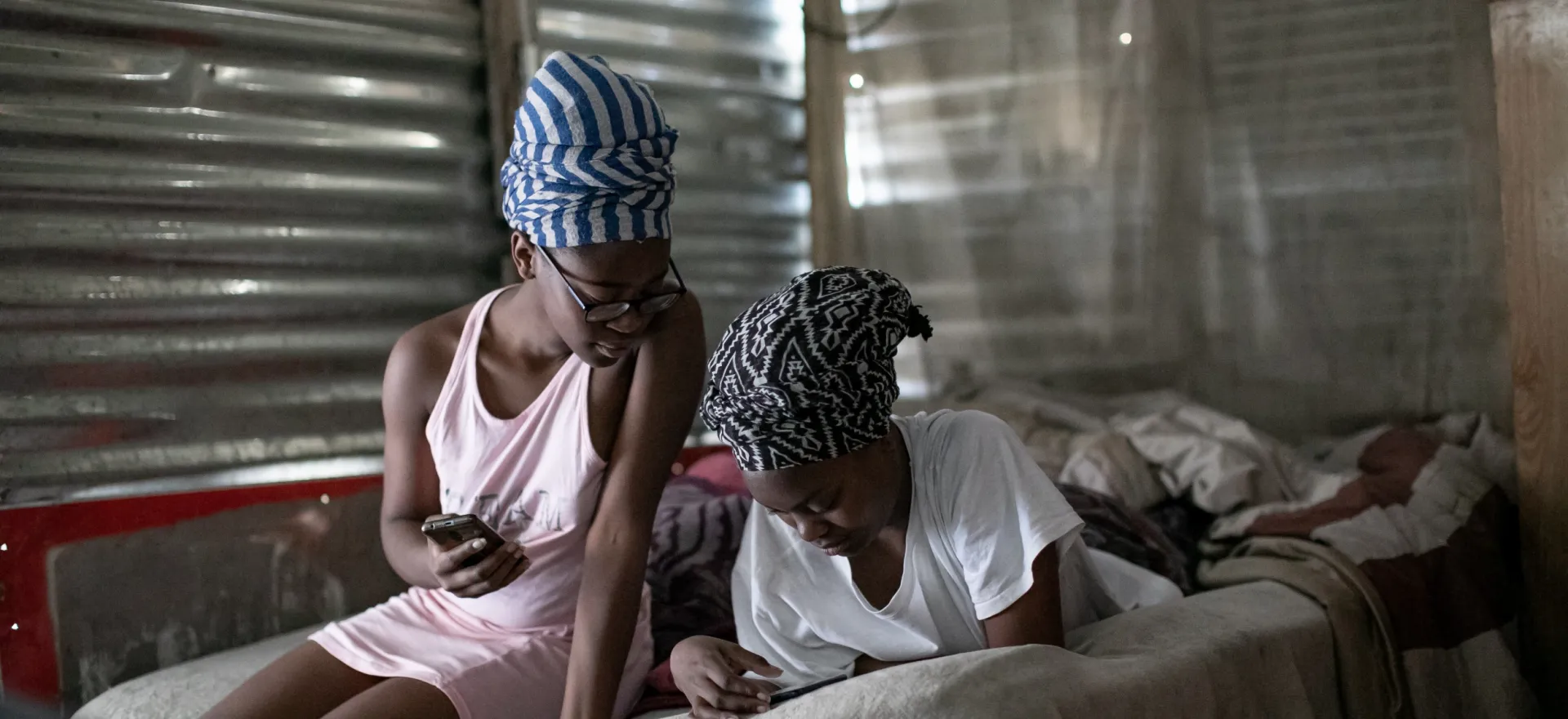 Two teenage girls, one lying down and the other sitting next to her on the bed in a sink house, look at their mobile phones.
