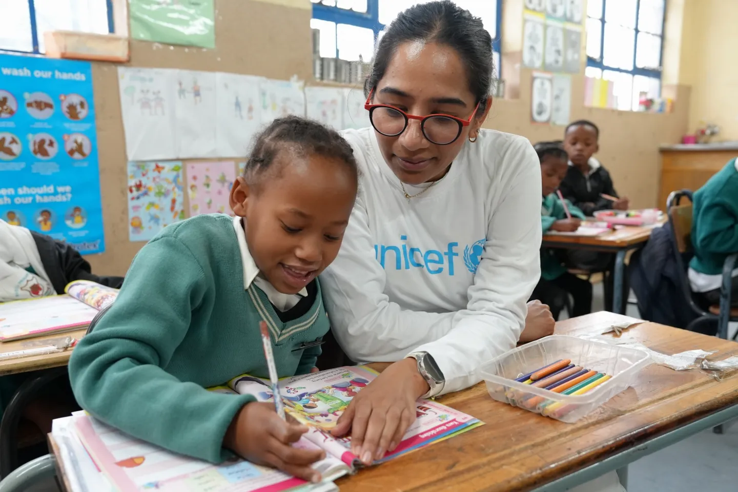 A girl in a green school uniform writes in a book on her desk in class, with a woman wearing a white sweatshirt with UNICEF on the front sits next to her.