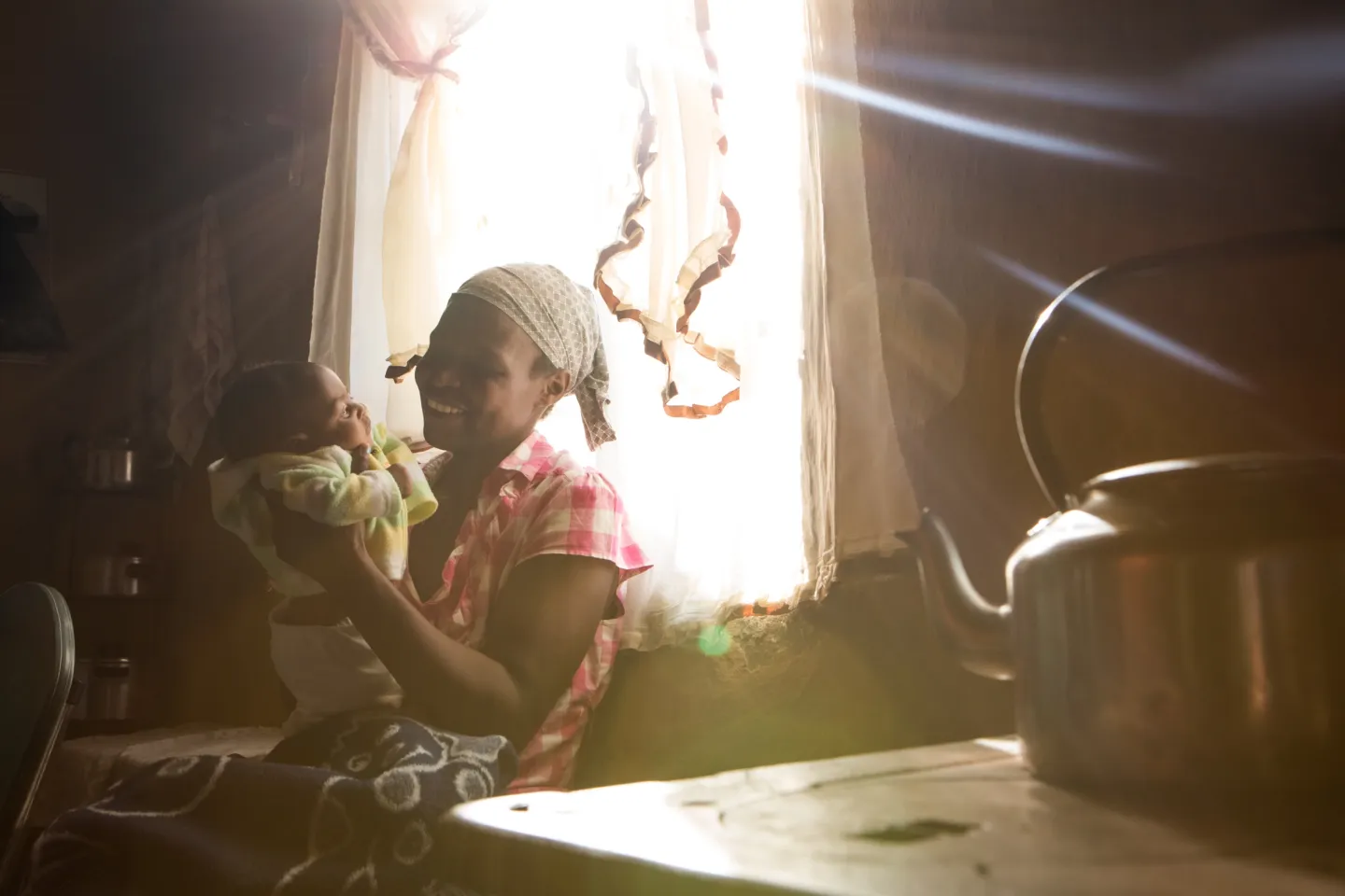 A woman sitting in front of a window holds her baby, with a kettle on a stove in the foreground