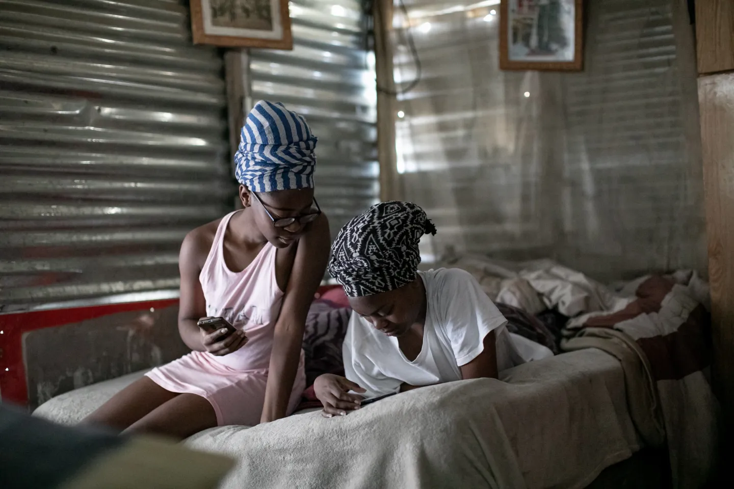 Two teenage girls, one lying down and the other sitting next to her on the bed in a sink house, look at their mobile phones.