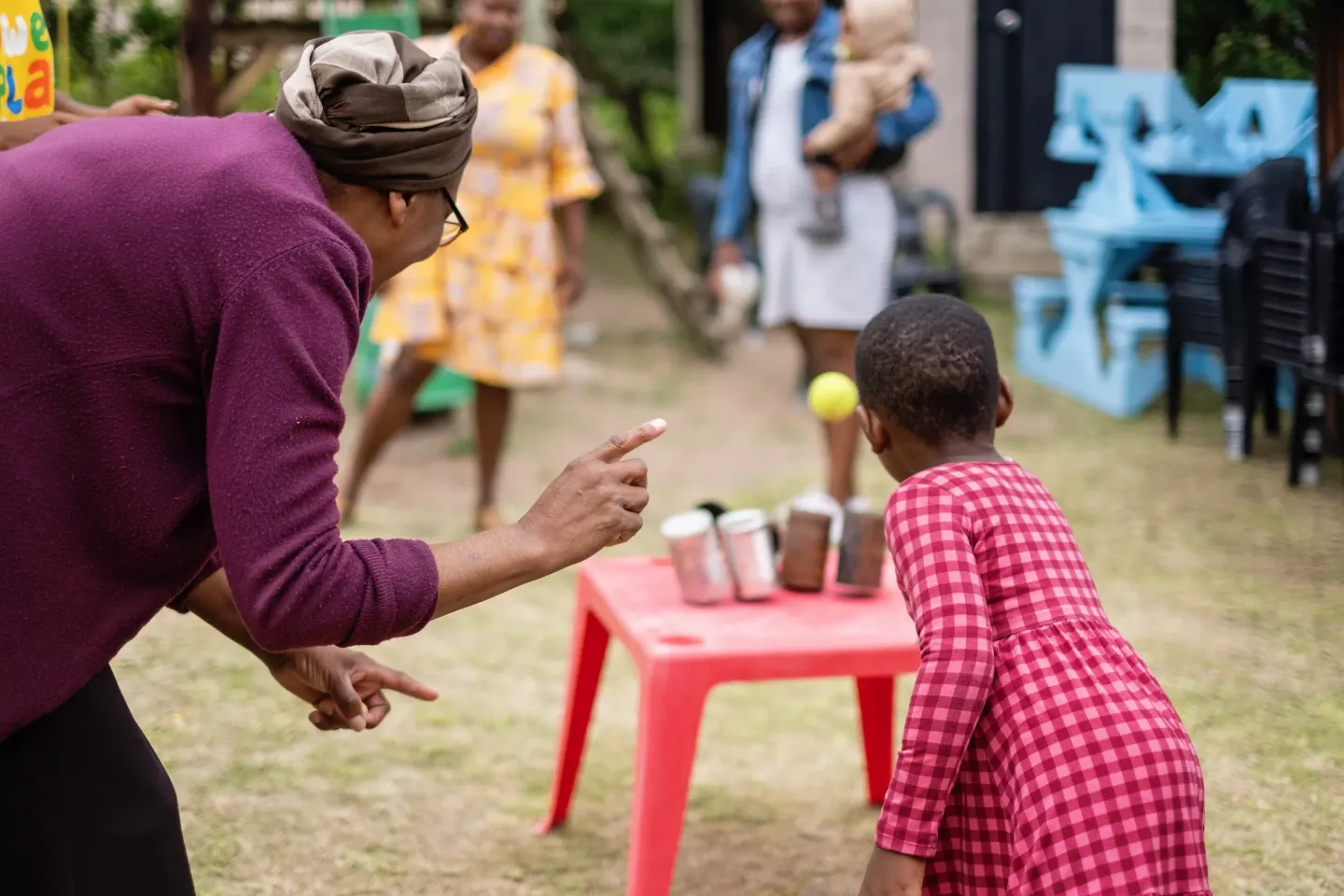 A little girl wearing a red dress threw a tennis ball towards at stacked cans on a table with her granny instructing her from the side.