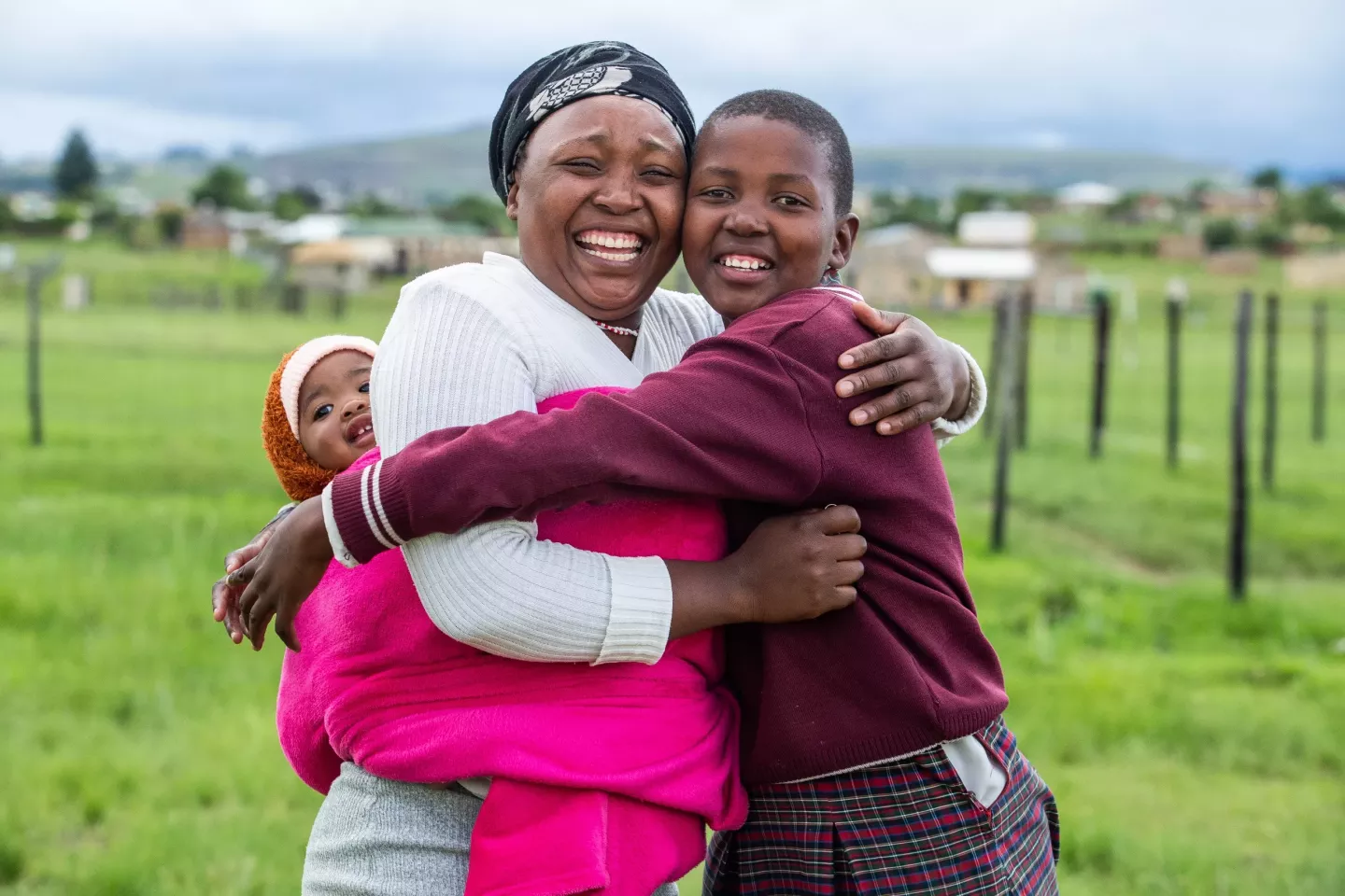 A woman carrying her infant daughter on her back, hugs her teenage daughter who is wearing a school uniform.