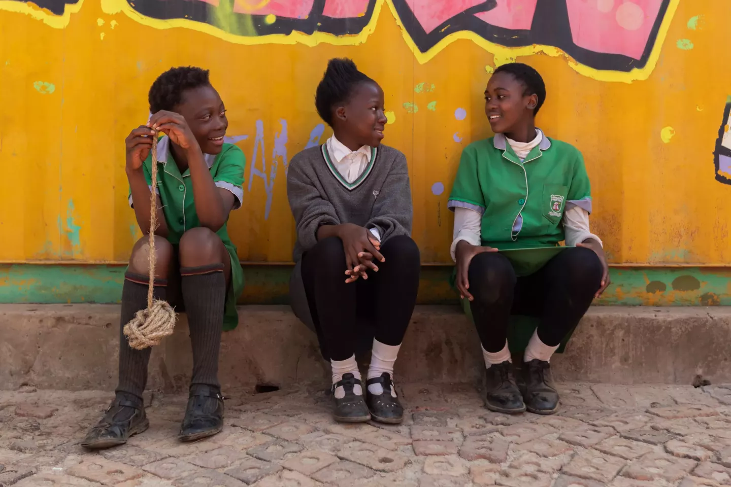 Three girls in school uniforms sits chatting in front of a a yellow wall.