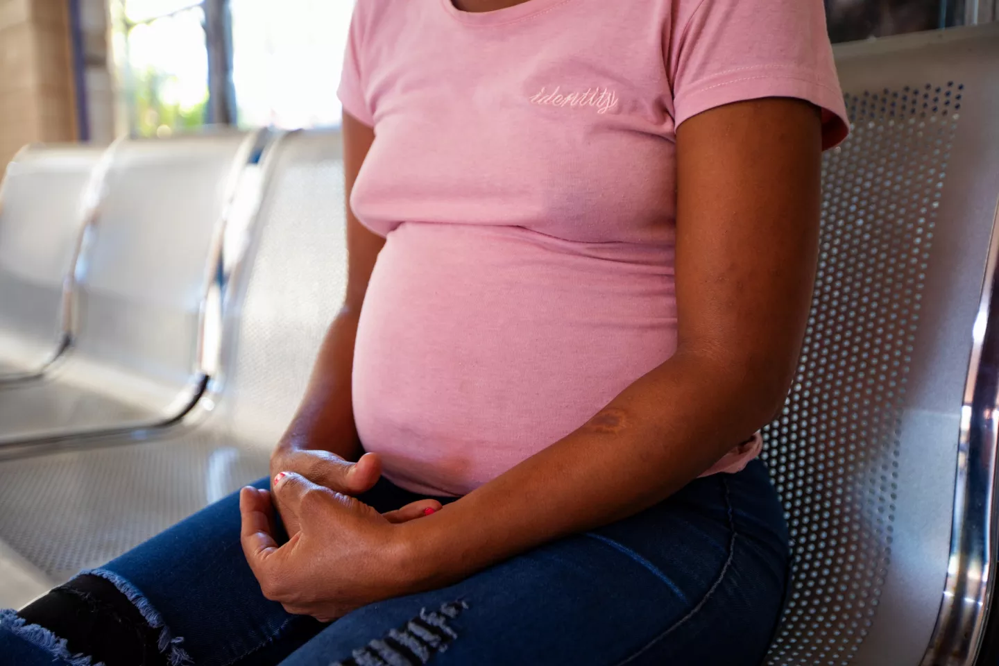 The torso of a pregnant woman wearing a pink t-shirt with jeans, sitting with her hands folded in her lap.