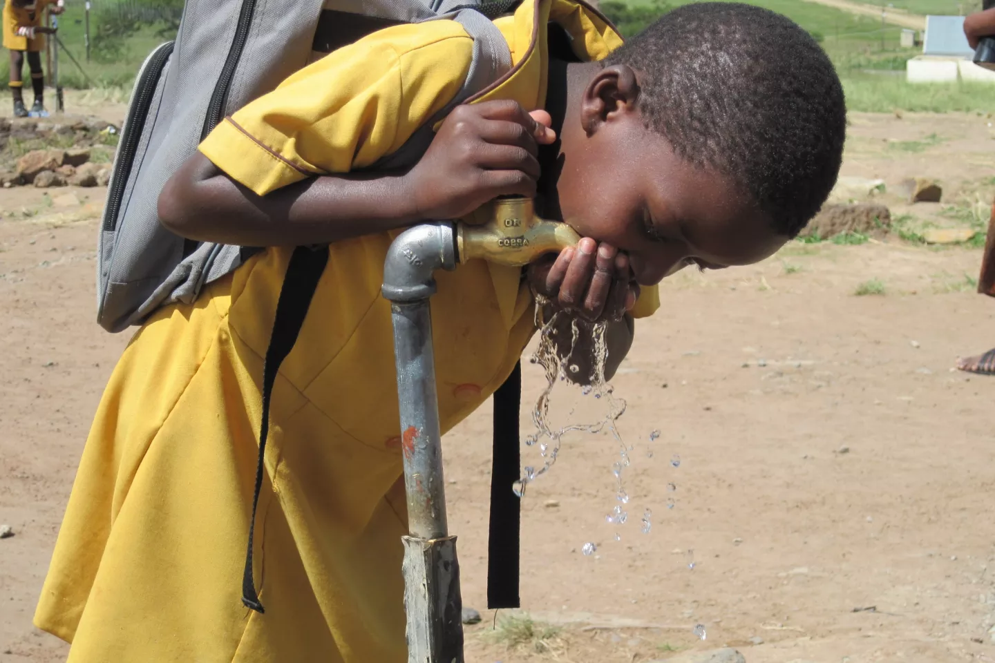A girl carrying a schoolbag on her back and wearing a yellow school uniform drinks water from a tap.