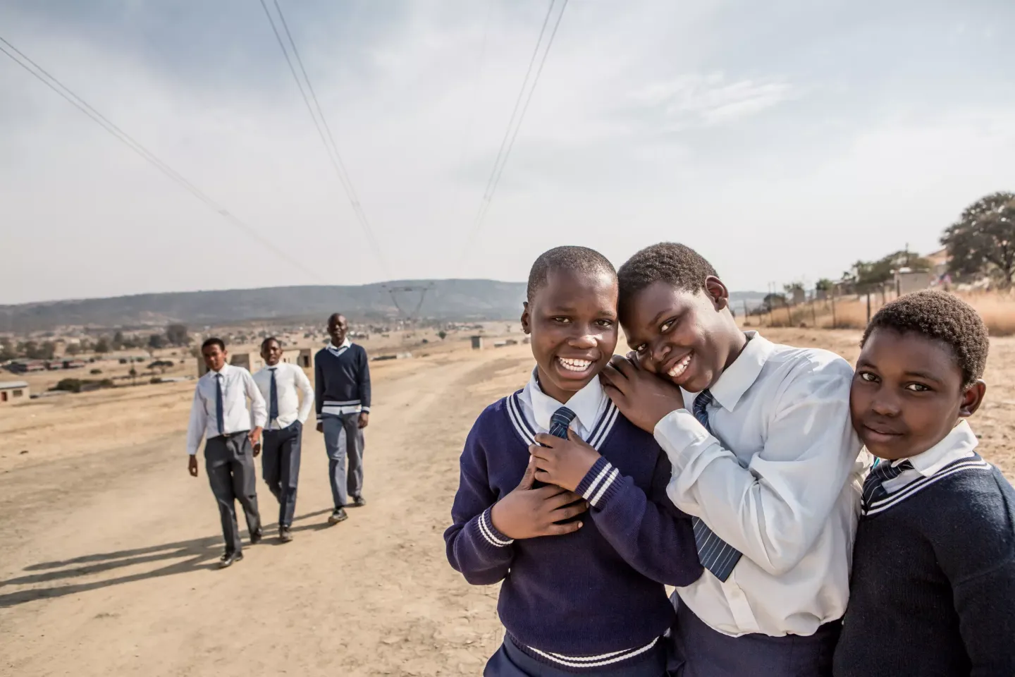Three teenage girls in school uniforms smile happily with a group of boys in school uniform walking some distance behind them on a dirt road.