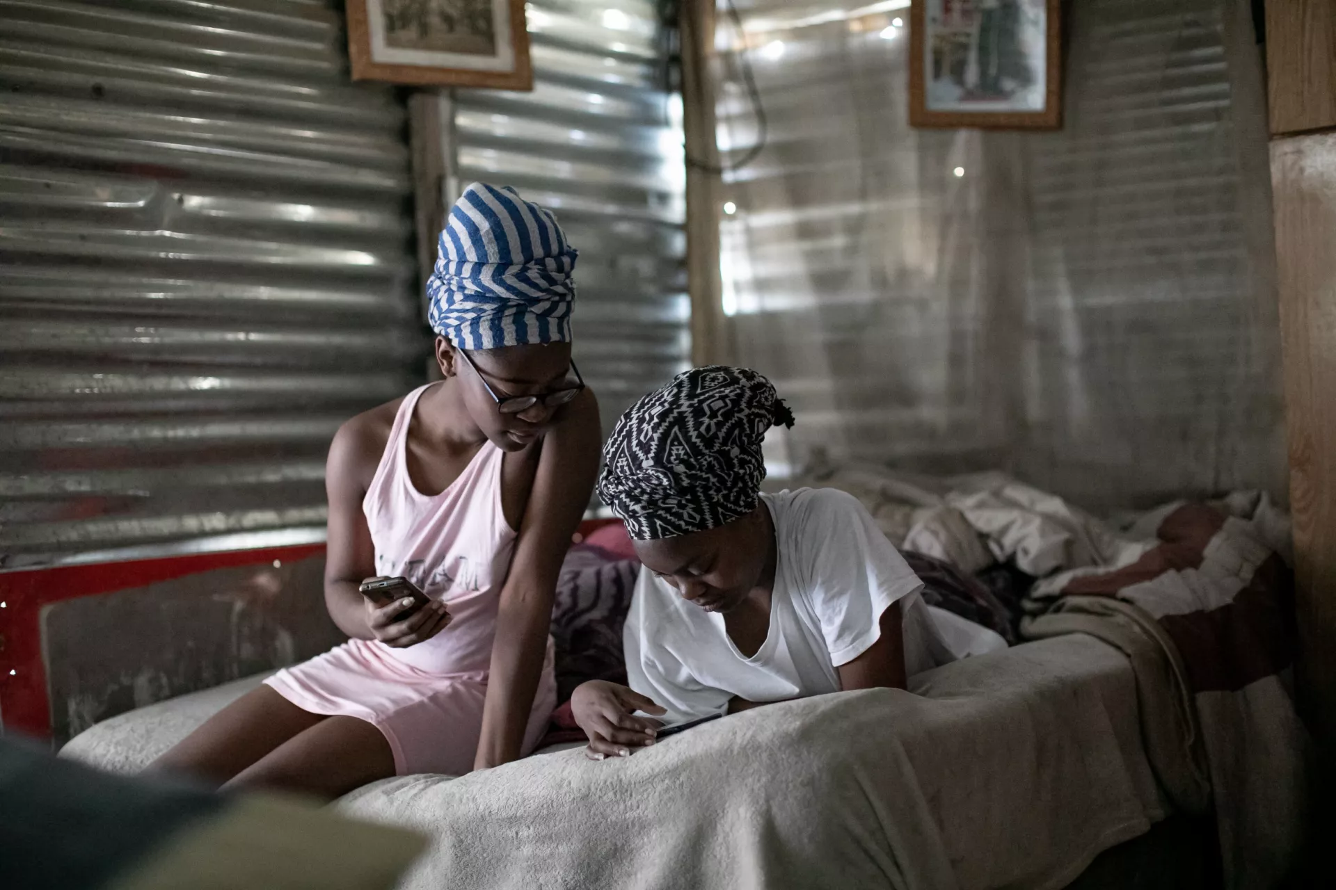 Two teenage girls, one lying down and the other sitting next to her on the bed in a sink house, look at their mobile phones.