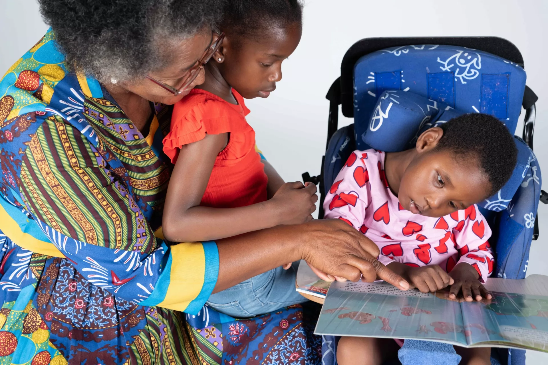 A granny with little girl on her lap plays with her sister who is disabled and sitting in a special chair next to them. 