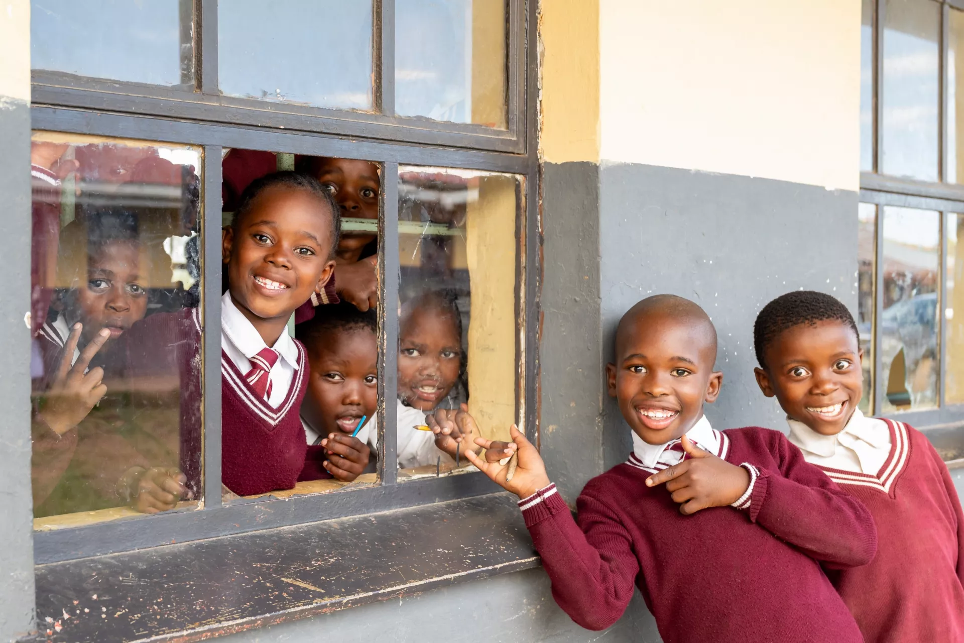 Primary school children peek through a broken classroom window, with their friends outside giving a thumbs up.