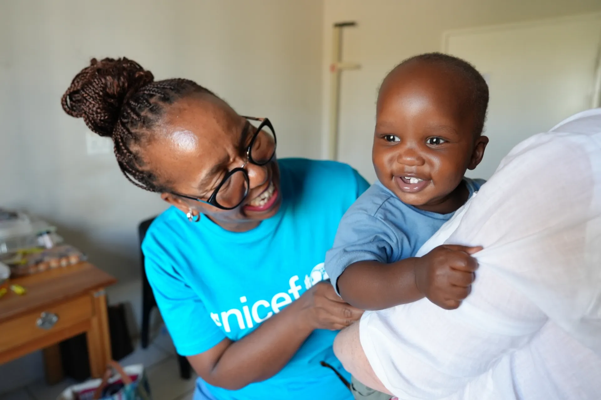 A UNICEF staff member in a blue UNICEF plays with a baby boy held by his mother.