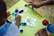 A pile of colourful bottlecaps are spread out on a green table. A little arm is visible, picking up a cap.
