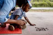 A little girl sits on the ground crossed-legged, leaning forward and looking at pebbles in a chalk-drawn square. Her dad in next to her giving her advice.