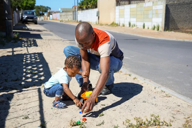 A father plays with his daughter on a sandy pavement outside their home.