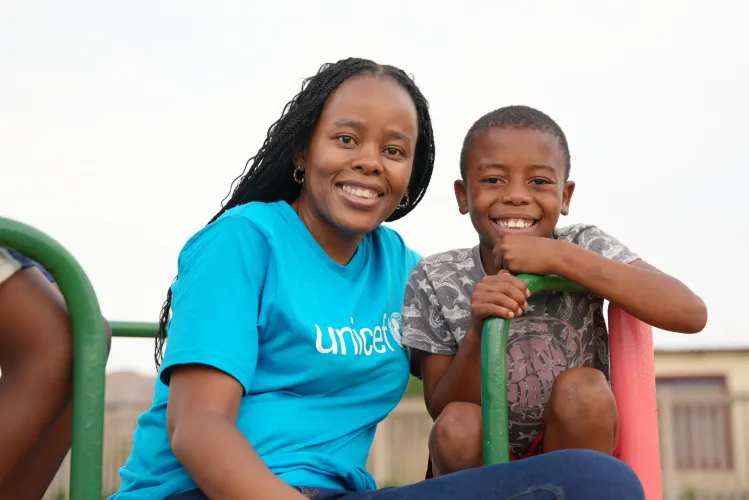 A UNICEF staff member wearing a blue t-shirt sits next to a young boy outside on playgound equipment.