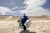 A woman carrying large bags of waste she's collected, walks across a bare stretch of land.
