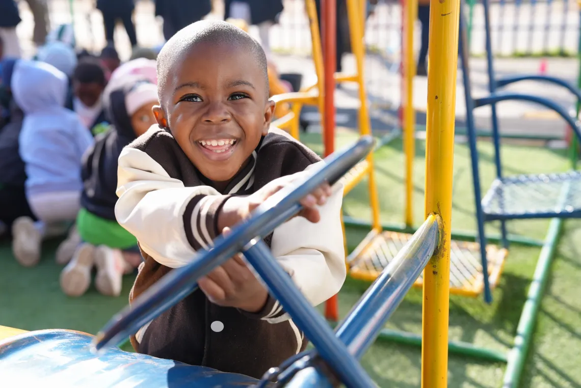 A little boy with his hands on a blue wheel, which is part of a larger colourful play installation, smiles happily.