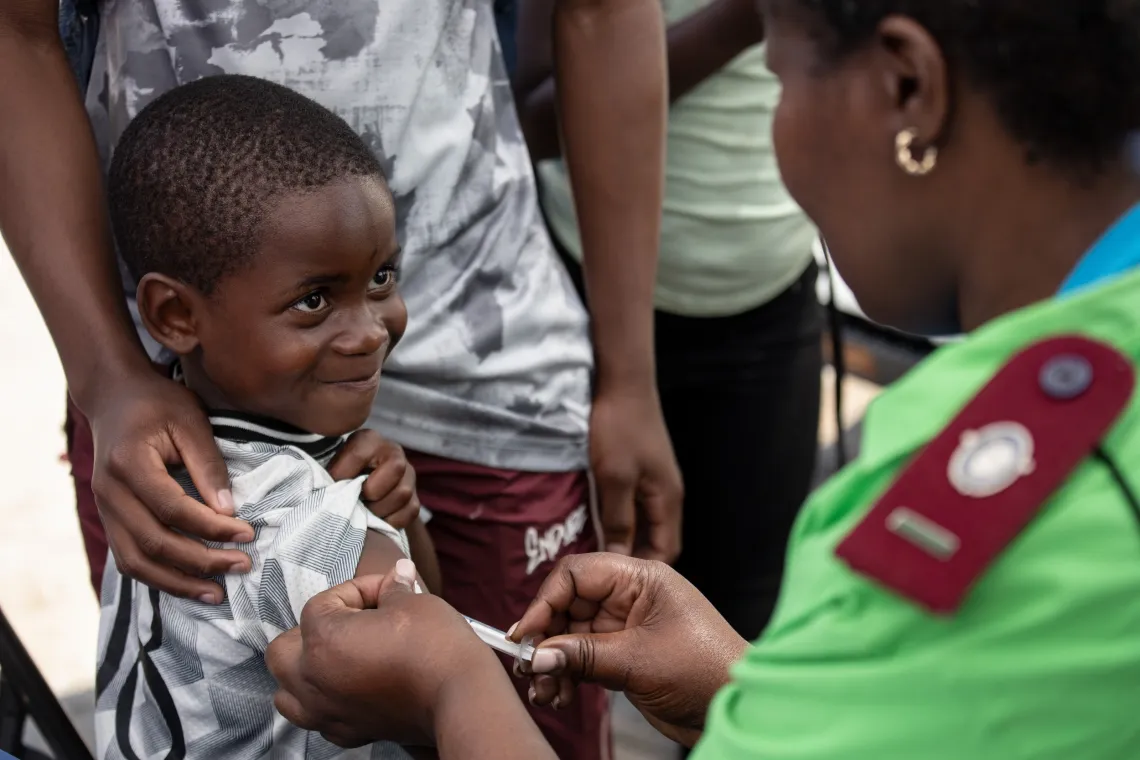 A little boy looks up smiling shyly, while a health worker wearing green gives him an injection in his arm. An older male wearing the same white and grey shirt as the little boy, hand is rested on the little boy's shoulder.