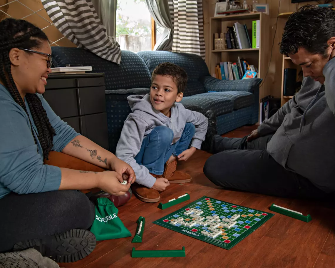 A smiling mother, father and son sitting on the floor in their living room with a scrabble board between them.