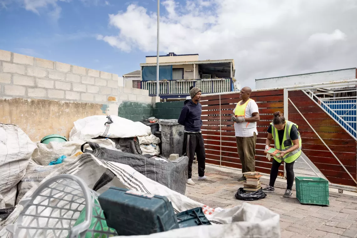 Two men and a woman stand next to large bags of recyclable waste.