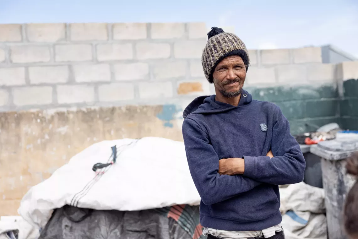 A man wearing a wool hat stands with folded arms in front of some waste that has been dropped off for recycling.