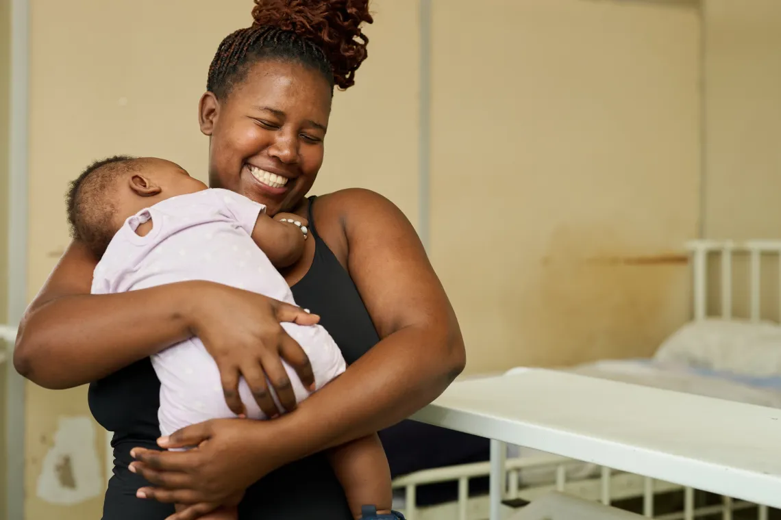 A young mother laughs as her baby girl in her arms, sleeps peacefully with her head on her shoulder.
