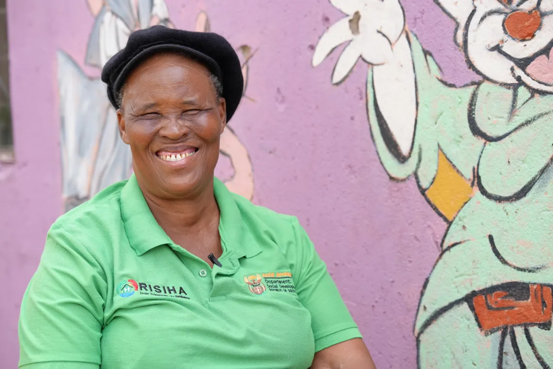 A woman wearing a green shirt and a black beret stands in front of a lilac painted wall.