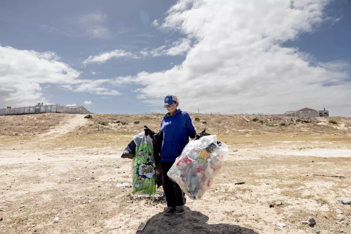 A woman carrying large bags of waste she's collected, walks across a bare stretch of land.