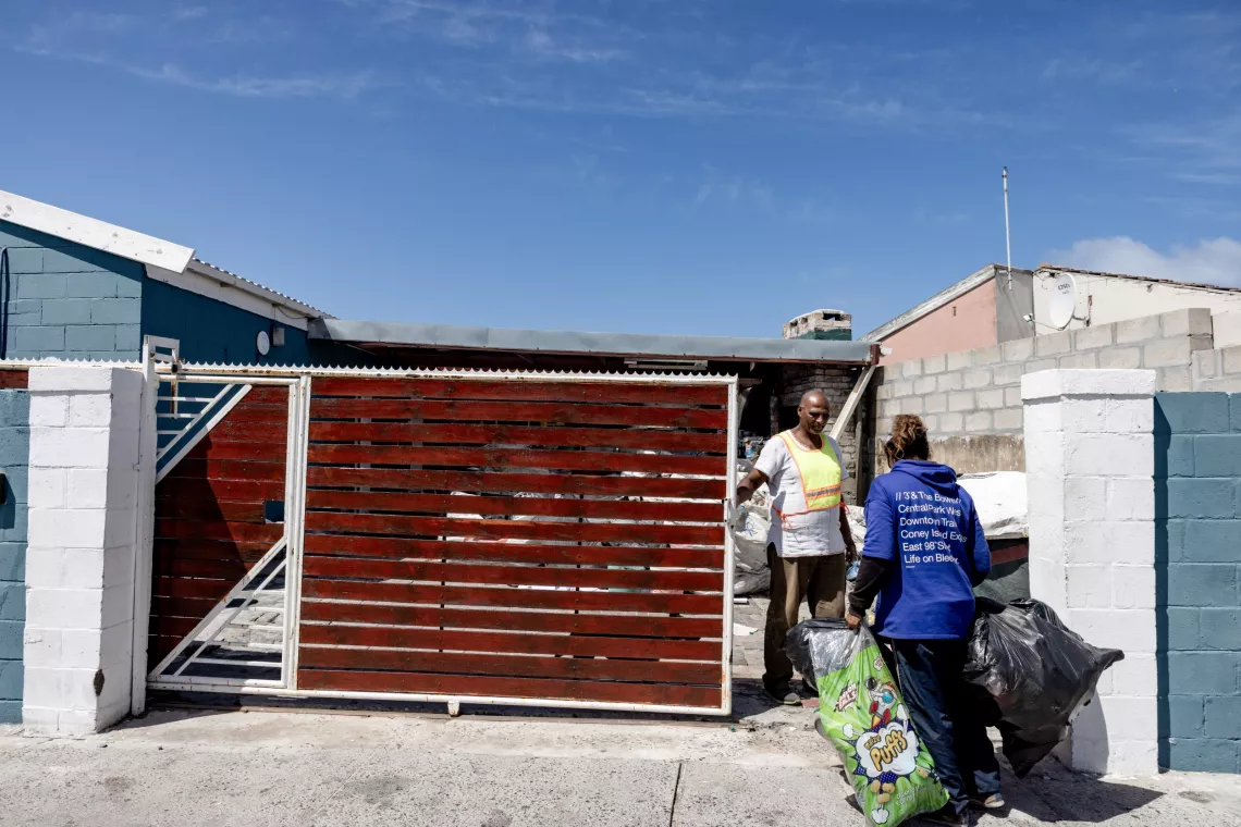A woman carries bags of collected recyclables up to a gate where a man is waiting for her.