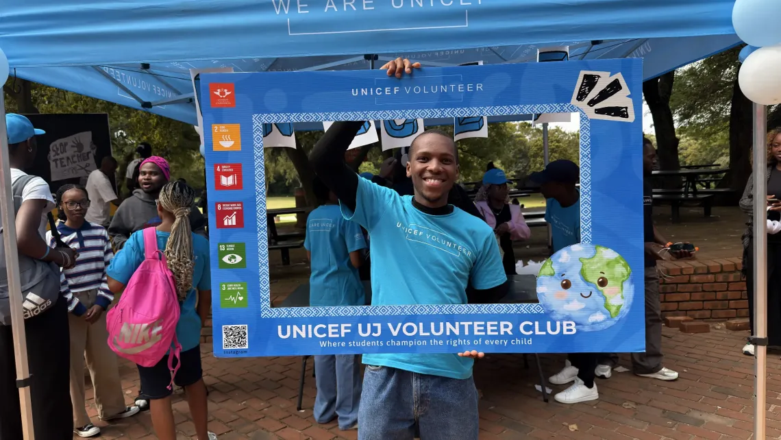 A male student wearing a blue UNICEF Volunteer t-shirt smile for a photo in a UNICEF Volunteer photo frame.
