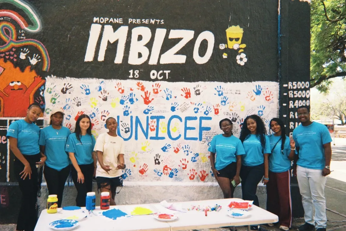 A group of students wearing blue UNICEF volunteer t-shirts pose for a photo in front of a large banner with colourful handprints on it.