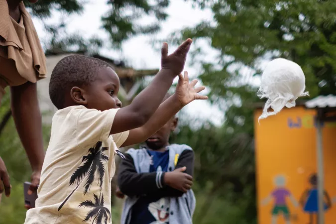 A ball made from plastic strips flies through the air from the outstretched arms of a little boy smiling happily.