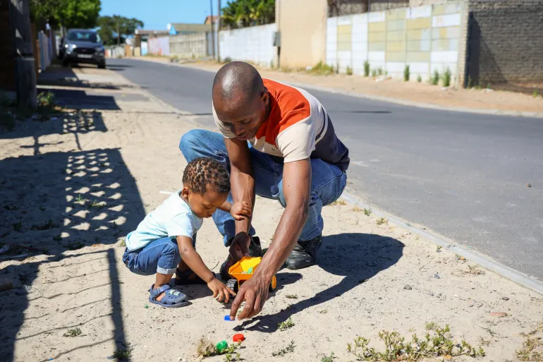 A father plays with his daughter on a sandy pavement outside their home.