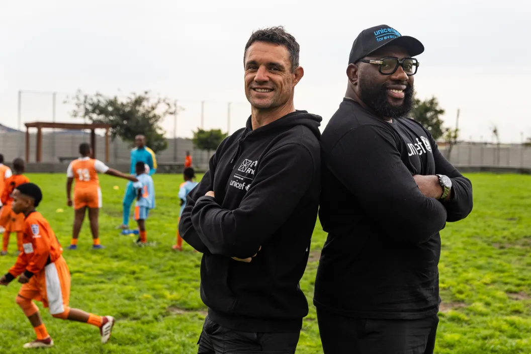 Retired rugby players Dan Carter from the New Zealand &ldquo;All Blacks&rdquo; and Tendai Mtawarira from the South African &ldquo;Springboks&rdquo; pose for a picture on a sports field with children playing in the background.