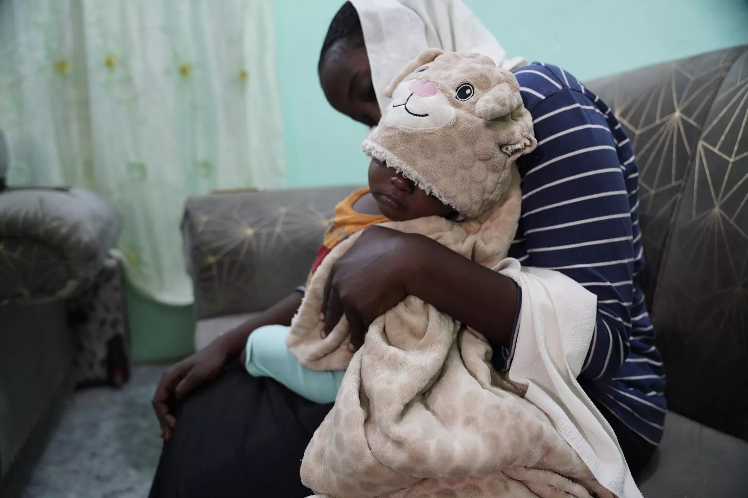 Sudan. A woman sits with her young son sleeping on her lap, wrapped in a blanket.