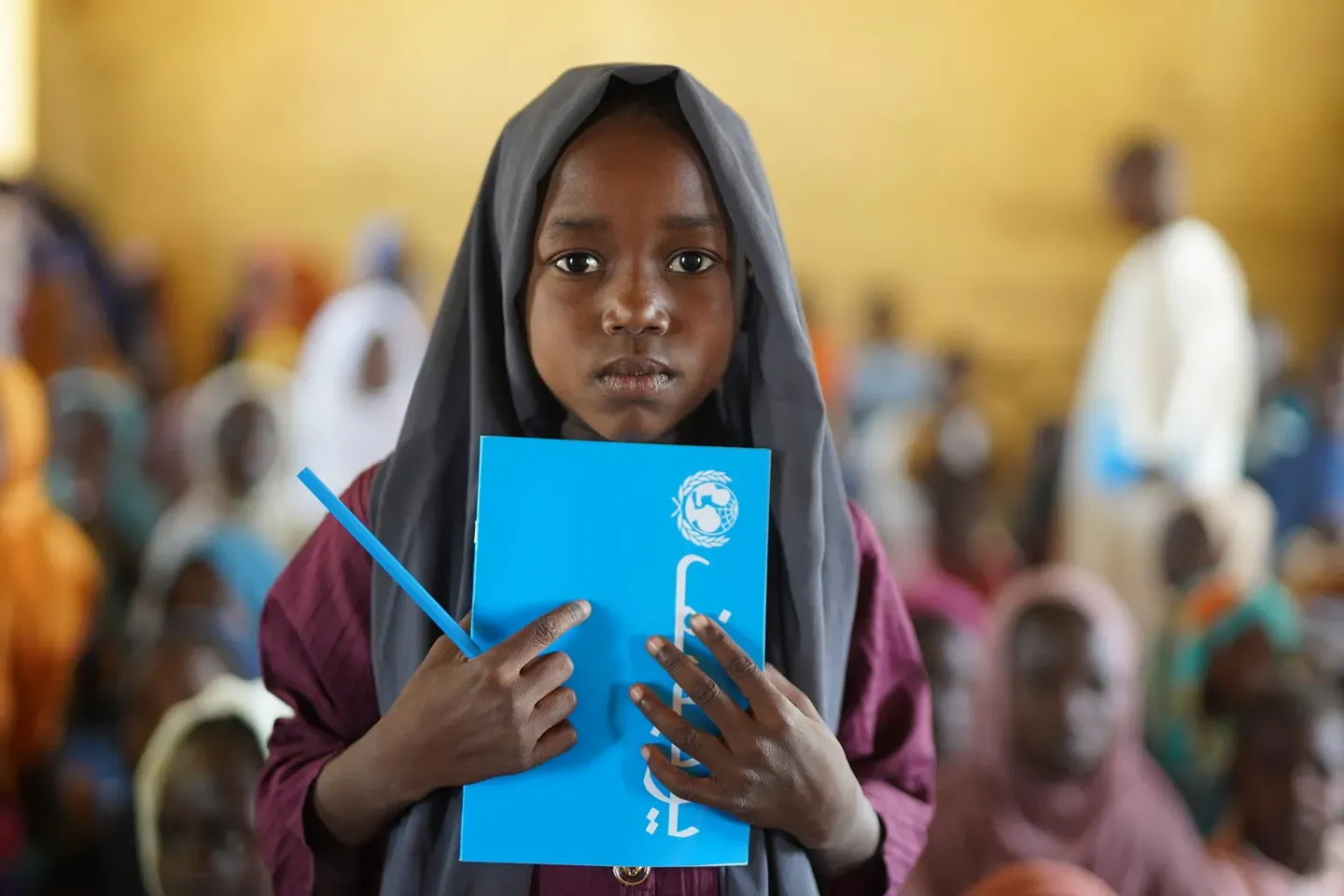 A child standing in a classroom while holding a UNICEF book and pencil 