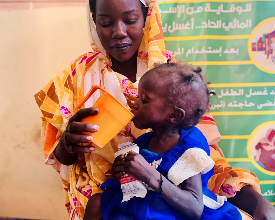 Sudanese woman gives child water from an orange cup.