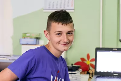 A smiling teenage boy is sitting by a desk with a laptop computer on it. 